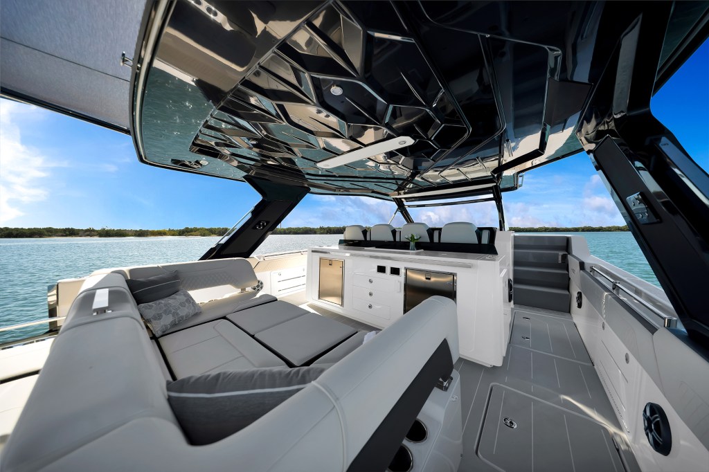 Spacious aft cockpit and bridge deck of an Aquila 45 Sport power catamaran, featuring a comfortable U-shaped outdoor seating lounge with grey cushions, a wet bar with stainless steel appliances, steps leading forward, and a dark geometric faceted hardtop underside design.
