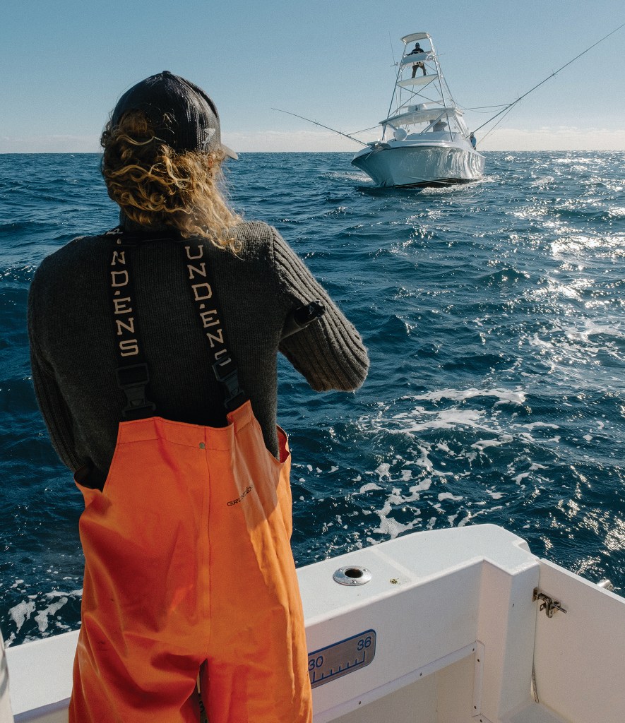 A person with long curly hair wearing orange fishing bib overalls stands on the deck of a boat, looking out at a sportfishing boat with a tuna tower out on open water.
