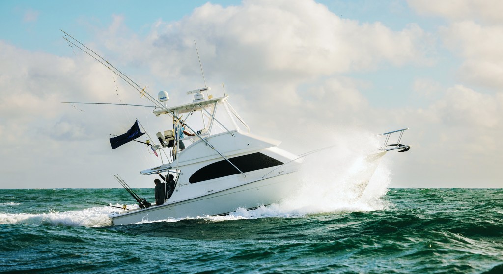 A white convertible sportfishing boat with outriggers and a flybridge cutting through choppy green ocean water, splashing spray from its bow under a partly cloudy sky.