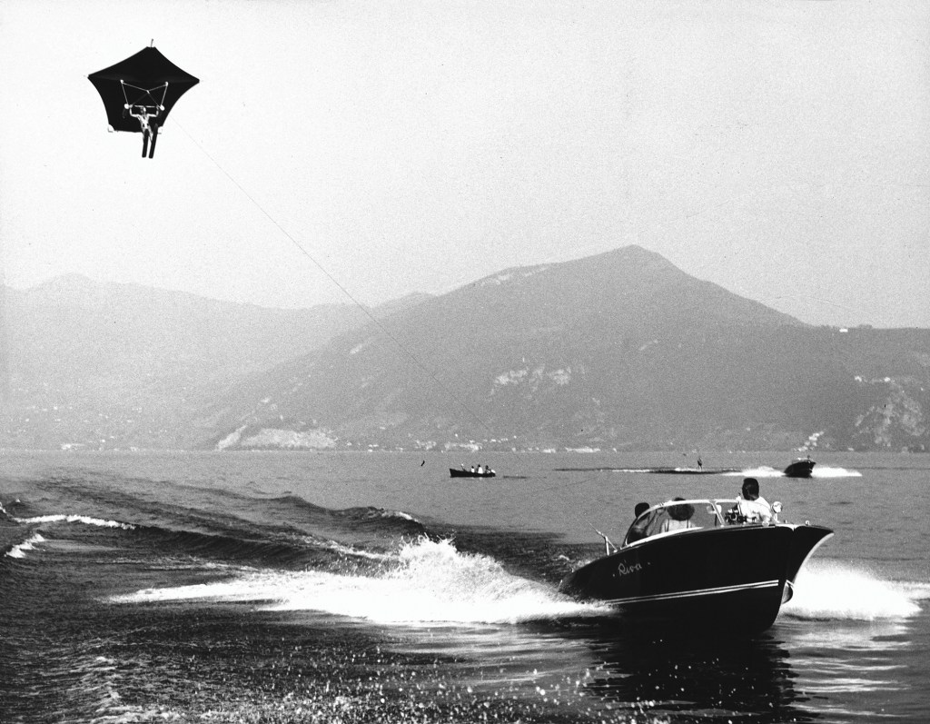 Vintage black-and-white photograph of a classic wooden Riva motorboat towing a parasailor across the water.