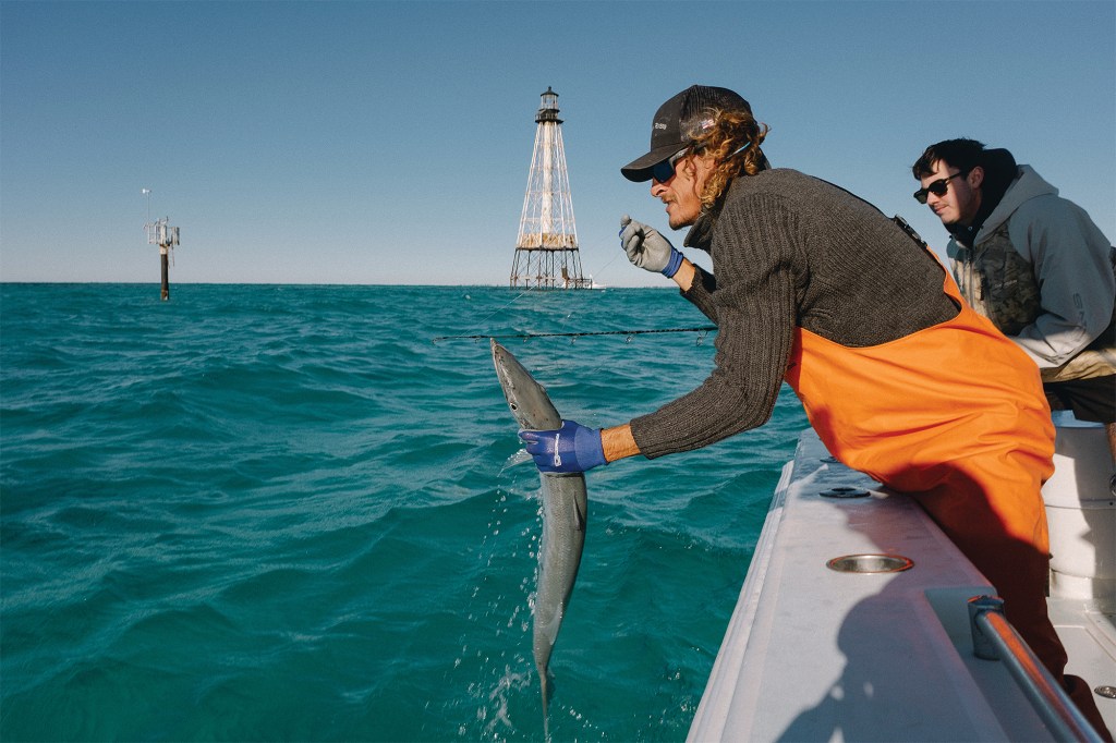 An angler wearing orange fishing bibs leans over the side of a boat to lift a long, slender fish out of the water, with a historic offshore lattice lighthouse visible in the background under a clear blue sky.