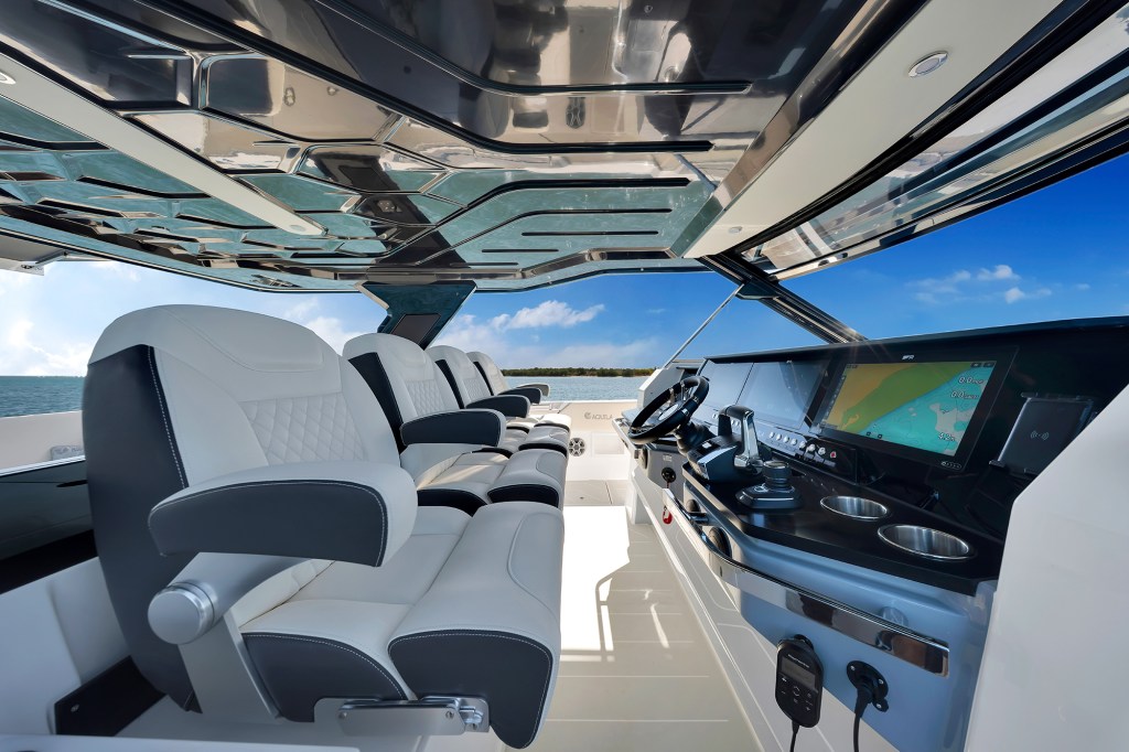 The helm station of an Aquila 45 Sport power catamaran featuring four captain's chairs under a hardtop, a row of large navigation display screens, steering wheel, and throttle controls.