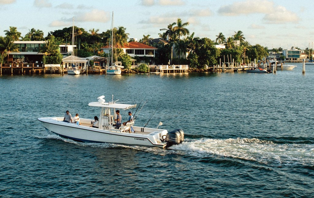A white Contender center console powerboat carrying several passengers cruises along a waterway, with lush palm trees, waterfront homes, and sailboat masts in the background.