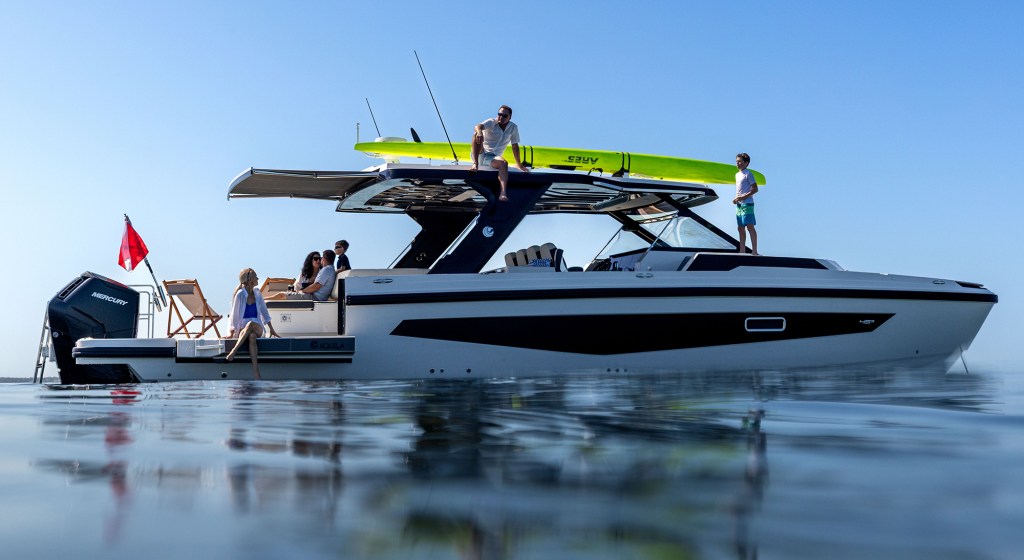 Side profile view of an Aquila 45 Sport power catamaran anchored on calm water, showcasing passengers relaxing across the deck, fold-down side terraces in the cockpit, a yellow paddleboard on the roof rack, and large Mercury outboard engines.