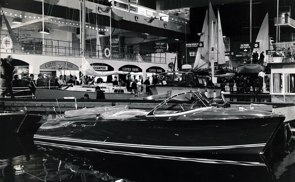 Vintage black-and-white photograph of a classic wooden Riva speedboat on display at a boat show.