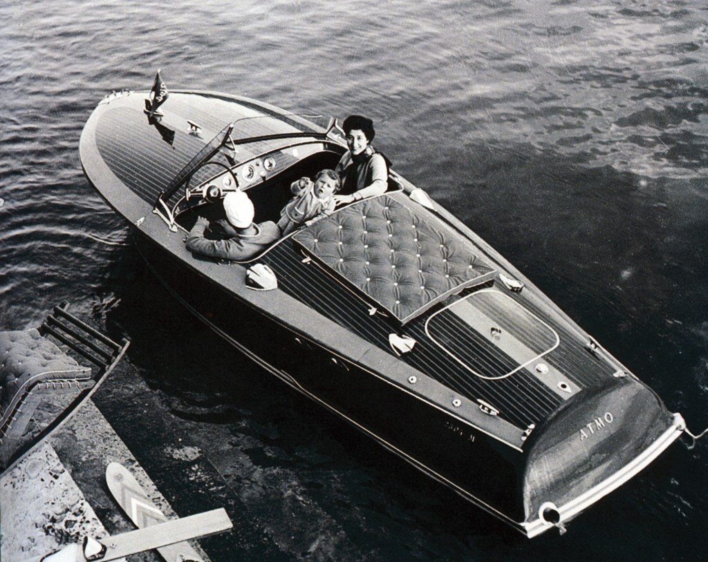 Vintage black-and-white aerial view of an early wooden Riva runabout motorboat carrying passengers near a dock.