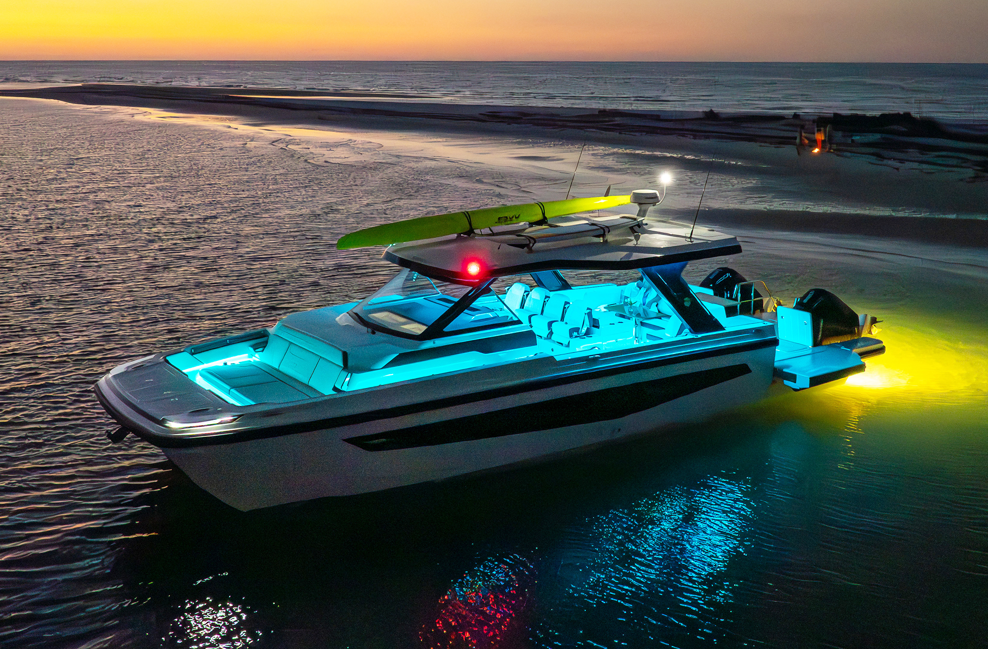 An aerial view of an Aquila 45 Sport power catamaran anchored near a shoreline at sunset, featuring blue hull LED accent lights, yellow underwater lights, twin Mercury outboards, and paddleboards secured to its hardtop roof rack.