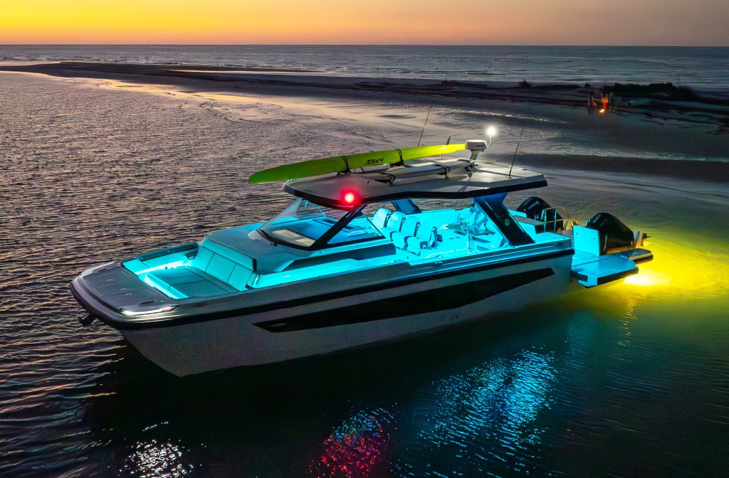An aerial view of an Aquila 45 Sport power catamaran anchored near a shoreline at sunset, featuring blue hull LED accent lights, yellow underwater lights, twin Mercury outboards, and paddleboards secured to its hardtop roof rack.