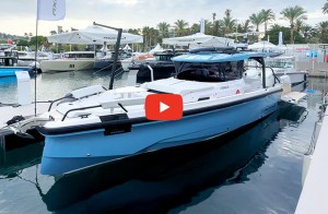 A light-blue and white Axopar motorboat docked at a marina during a boat show, surrounded by other vessels and palm trees under an overcast sky. A red play button icon is overlaid in the center of the image.