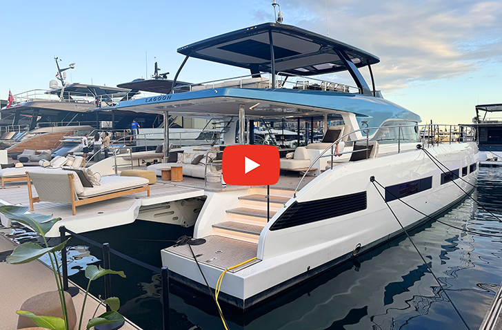 A luxury white and blue motor yacht docked at a marina, featuring a spacious open-air aft deck with modern lounge seating, an upper flybridge deck, steps leading down to the water, and a red video play icon centered in the frame.