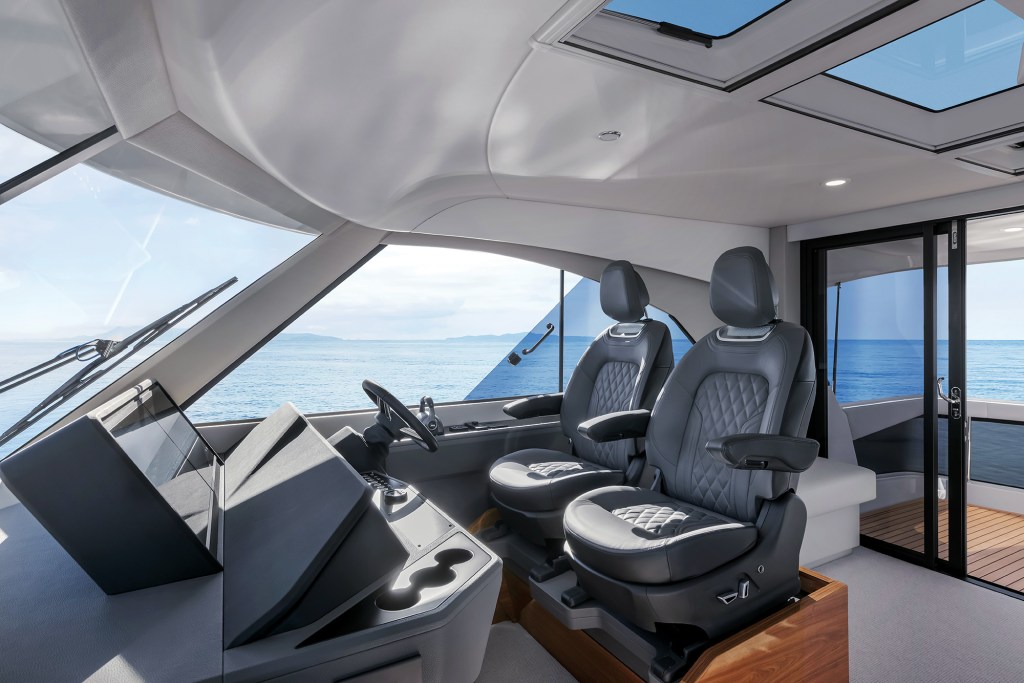 The interior helm station of a Maritimo M50 featuring twin dark leather captain's chairs, multi-function displays, overhead skylights, and large panoramic windows overlooking the ocean.