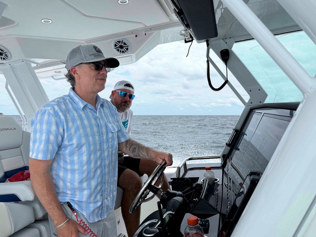 A captain stands at the covered helm station of a Formula center console boat holding the stainless steel wheel while driving on open water.