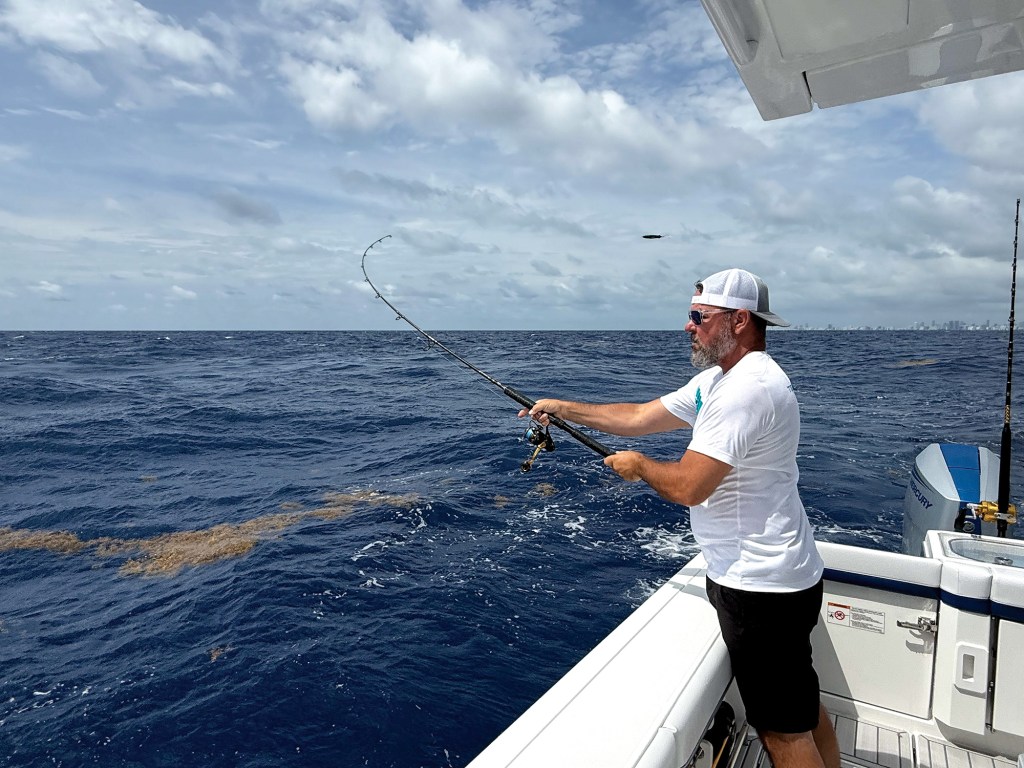 An angler standing on the gunwale of a Formula center console boat actively reeling in a fish with a bent fishing rod over deep ocean water.