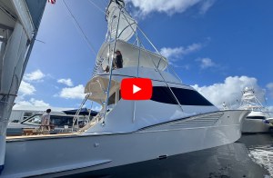 Dockside profile view of a white Hatteras 77 sportfishing yacht with a towering tuna tower and a red play button overlay at FLIBS.
