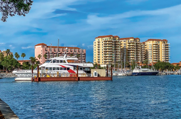 A white passenger ferry docked in a marina along the St. Petersburg, Florida waterfront with high-rise buildings and palm trees in the background.